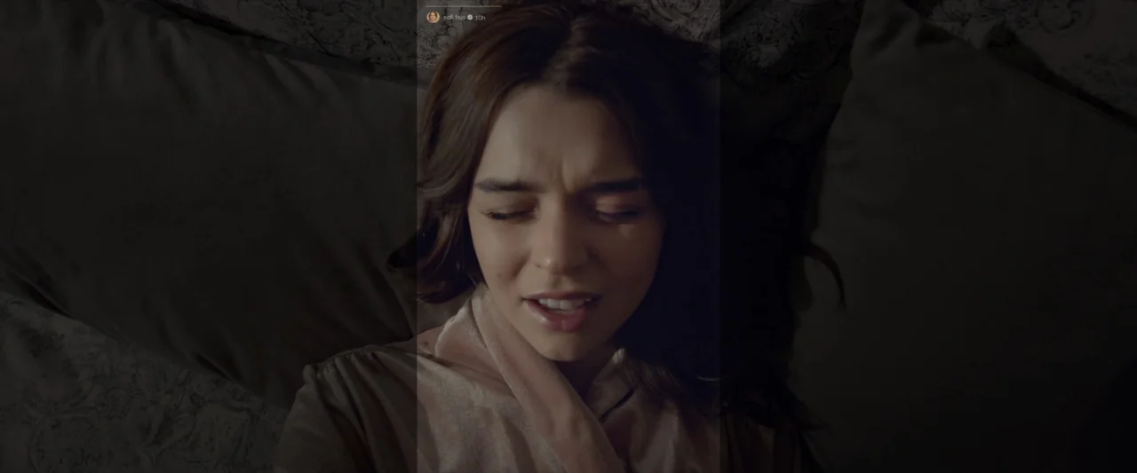 Like or Die (2023) movie still: closeup — A dimly lit bedroom features a woman resting on soft pillows against a dark headboard. The person lies…