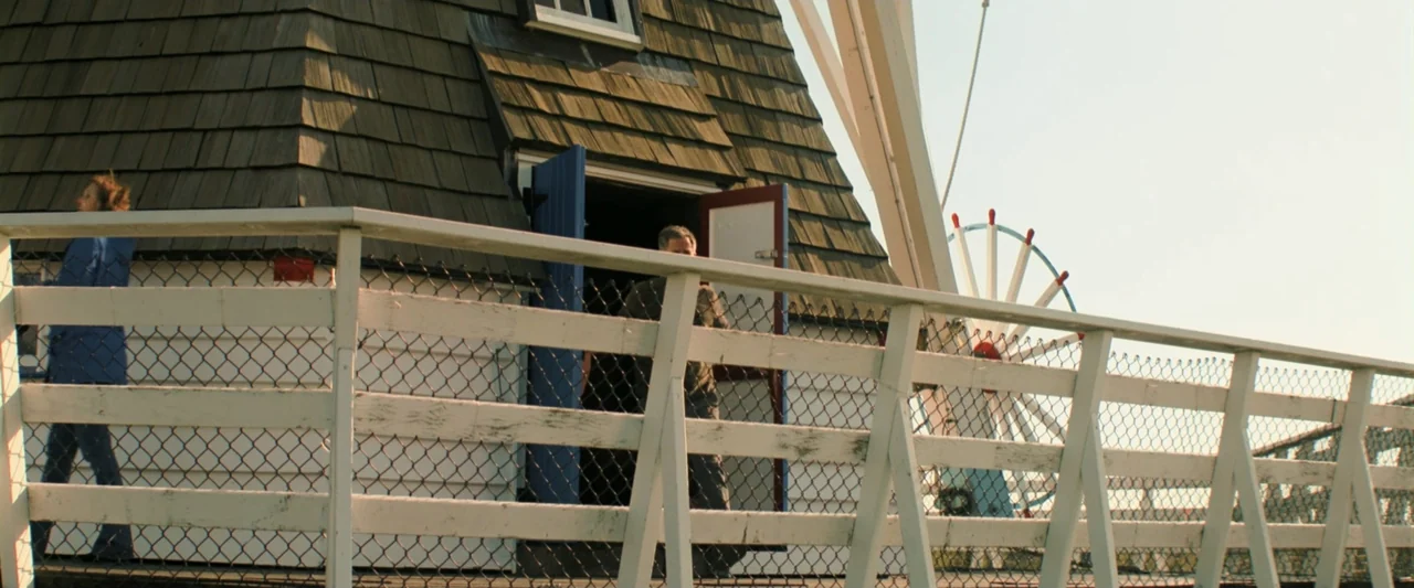 Holland (2025) movie still: wide — A wooden windmill structure stands beside a white railing against a clear sky. A man stands in the doorway while…