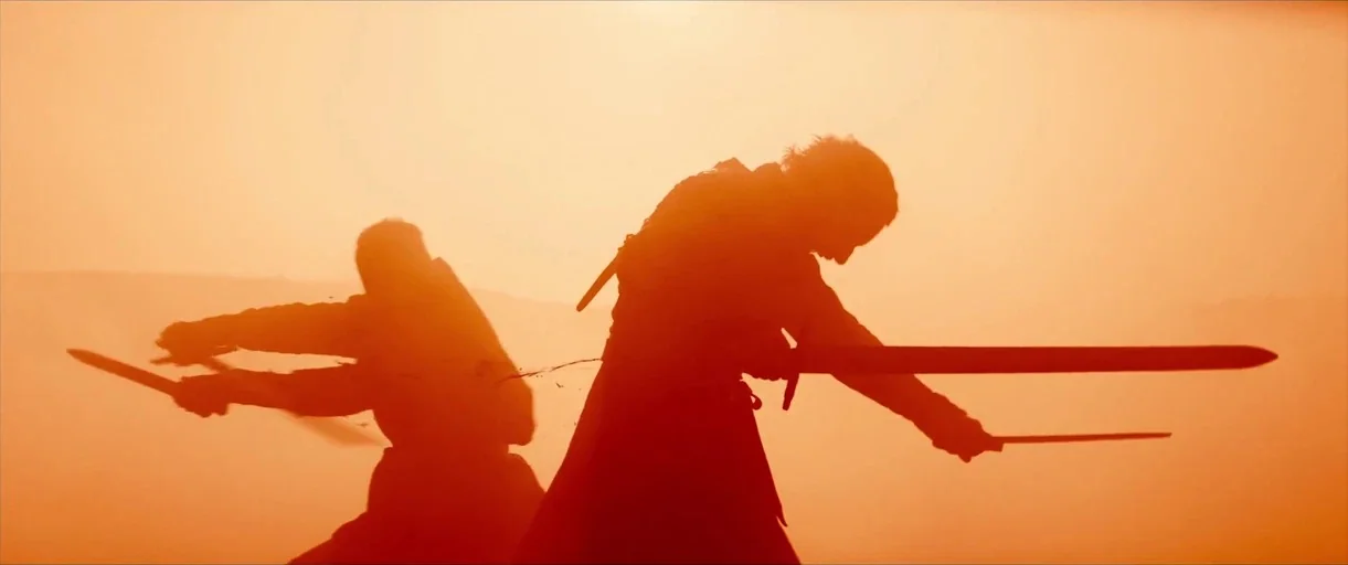 Macbeth (2015) movie still: medium — A dusty outdoor field lit by bright, hazy sunlight. Two silhouettes of armed warriors engage in sword combat…