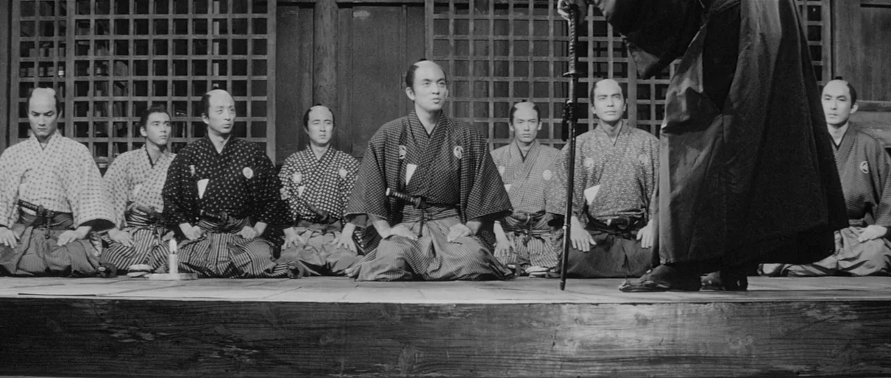Sanjuro (1962) movie still: fullbody — A group of men in traditional Japanese clothing kneel on a wooden floor. They face a figure standing with a…
