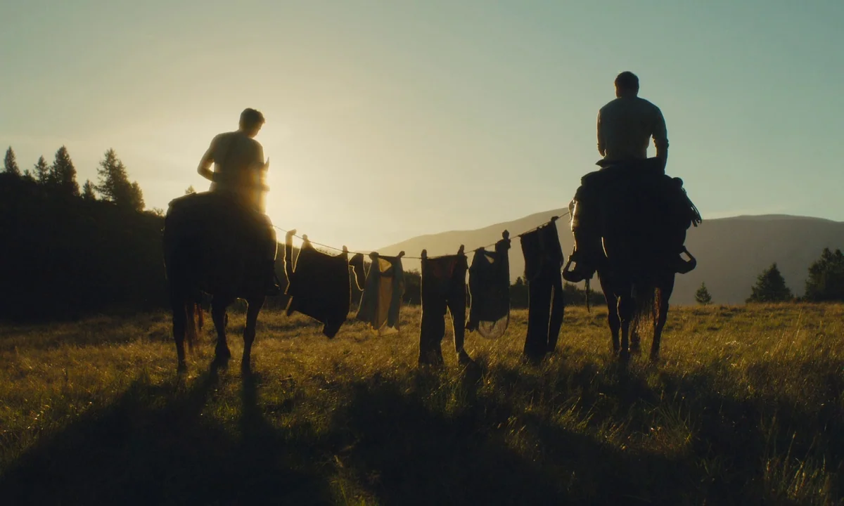 Slow West (2015) movie still: wide — An open mountain meadow at golden hour with distant hills under a clear sky. Two men sit on horseback, facing…