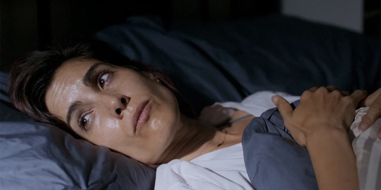 Demonic (2015) movie still: closeup — A dimly lit bedroom features a woman resting in bed under dark grey sheets. The woman lies still on her pillow…