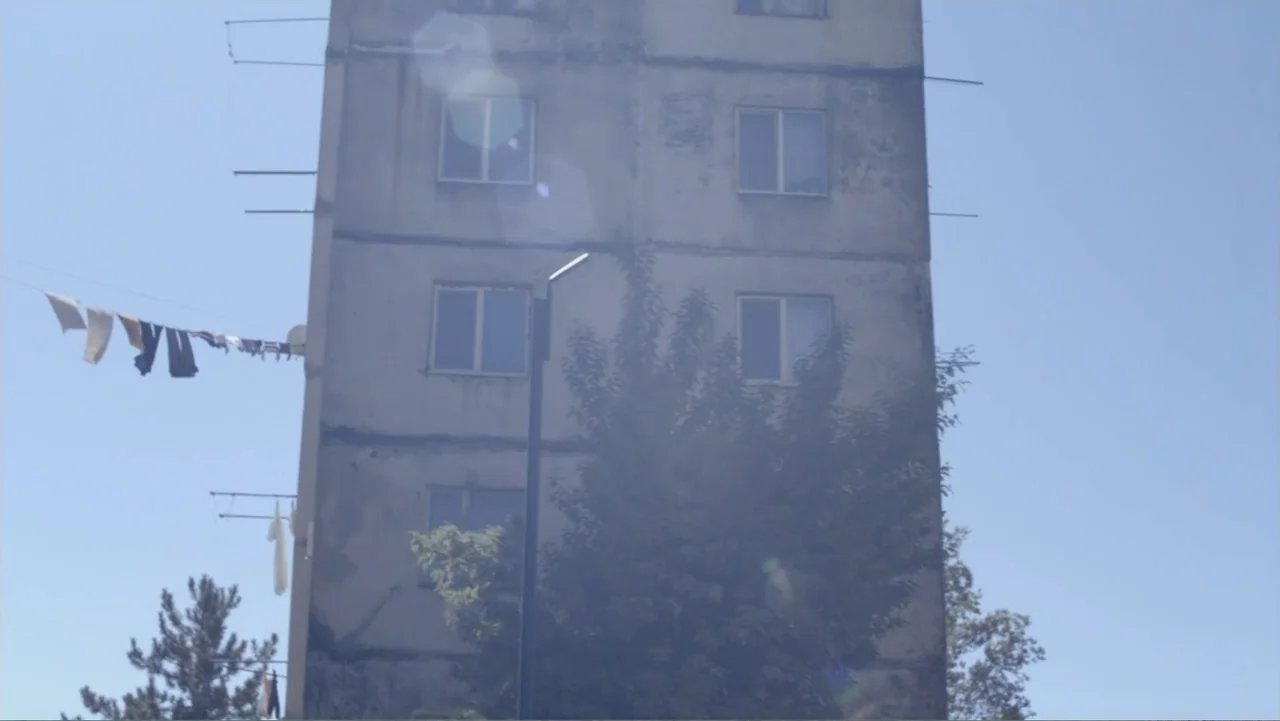 Letters to Max (2014) movie still: establishing — A grey multi-story apartment building with visible weathering and several windows is shown…