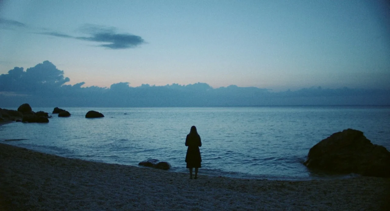Blow The Man Down (2019) movie still: wide — A sandy shoreline meets calm dark water under a blue dusk sky. A lone figure stands facing the ocean…