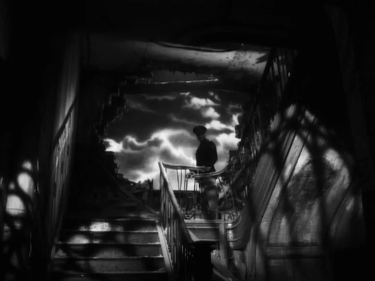 The Good German movie still: wide — A dark, dilapidated staircase features a massive hole in the exterior wall revealing clouds. A lone soldier…