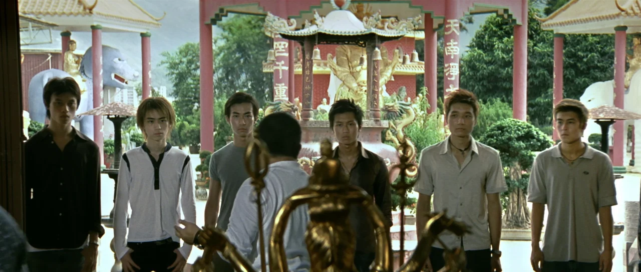 Infernal Affairs (2002) movie still: wide — A temple courtyard features ornate red columns and golden religious statues. Six young men stand in a…