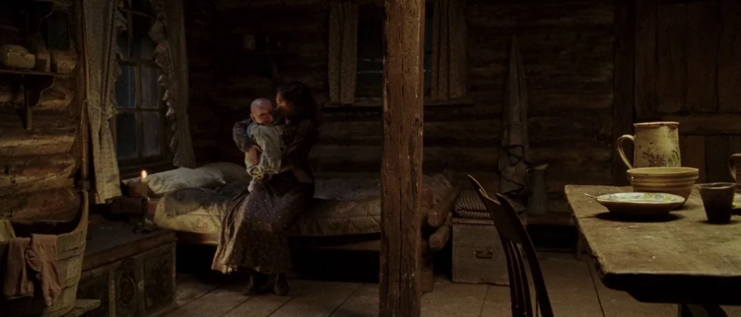 Cold Mountain (2003) movie still: wide — A dark log cabin interior features rough-hewn timber walls and a rustic wooden table. A woman holding an…