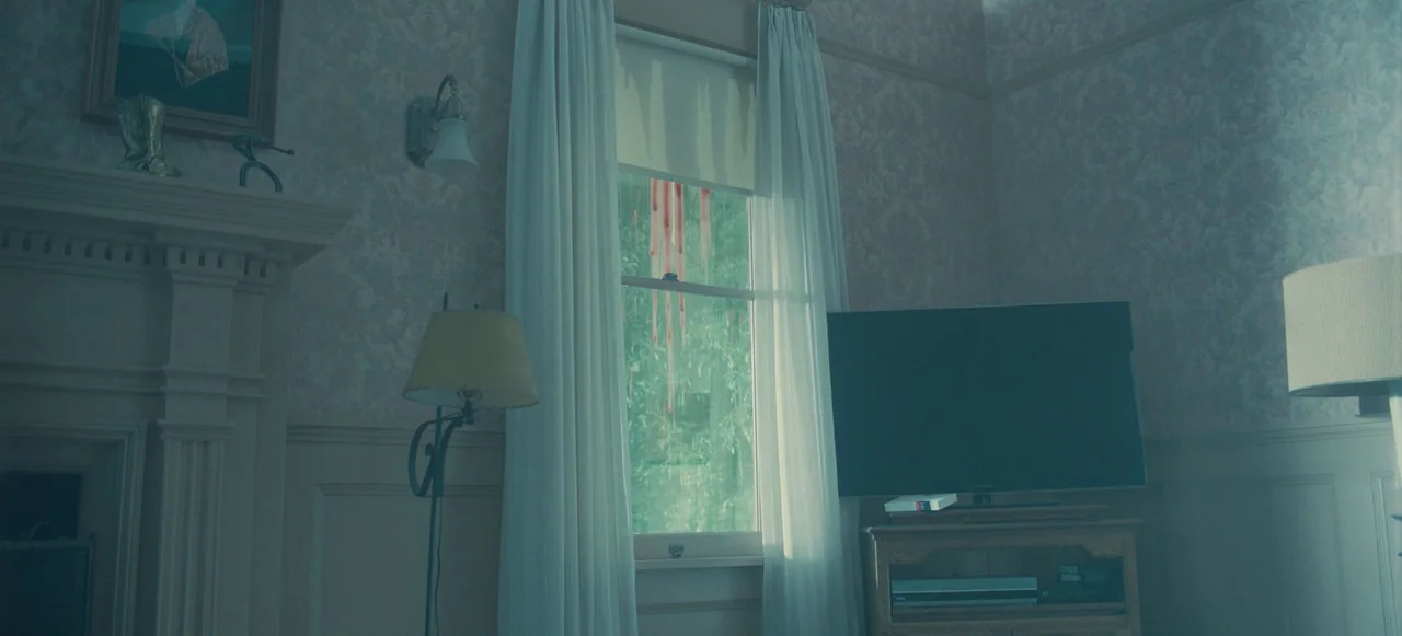 Nope (2022) movie still: wide — A living room interior with patterned wallpaper features a window covered in streaks of blood. The room contains a…