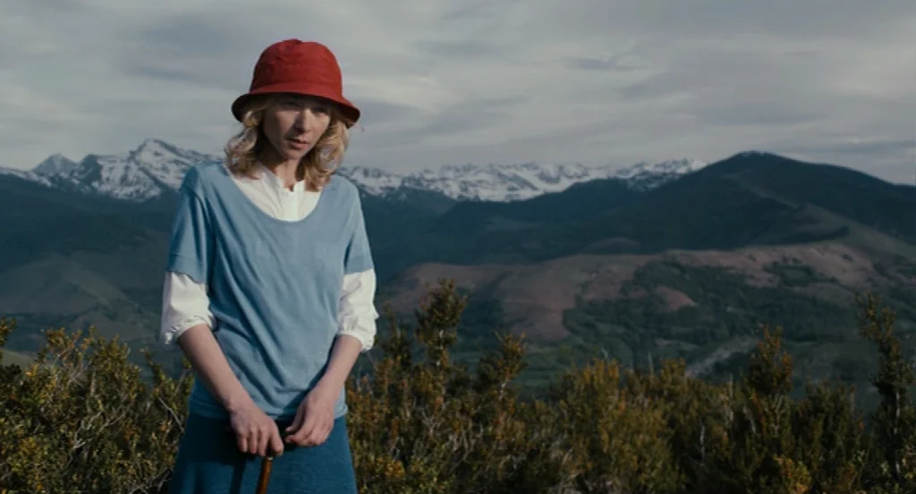 Lourdes movie still: medium — A woman stands on a grassy mountain ridge overlooking a range of snow-capped peaks. She wears a red hat and blue…