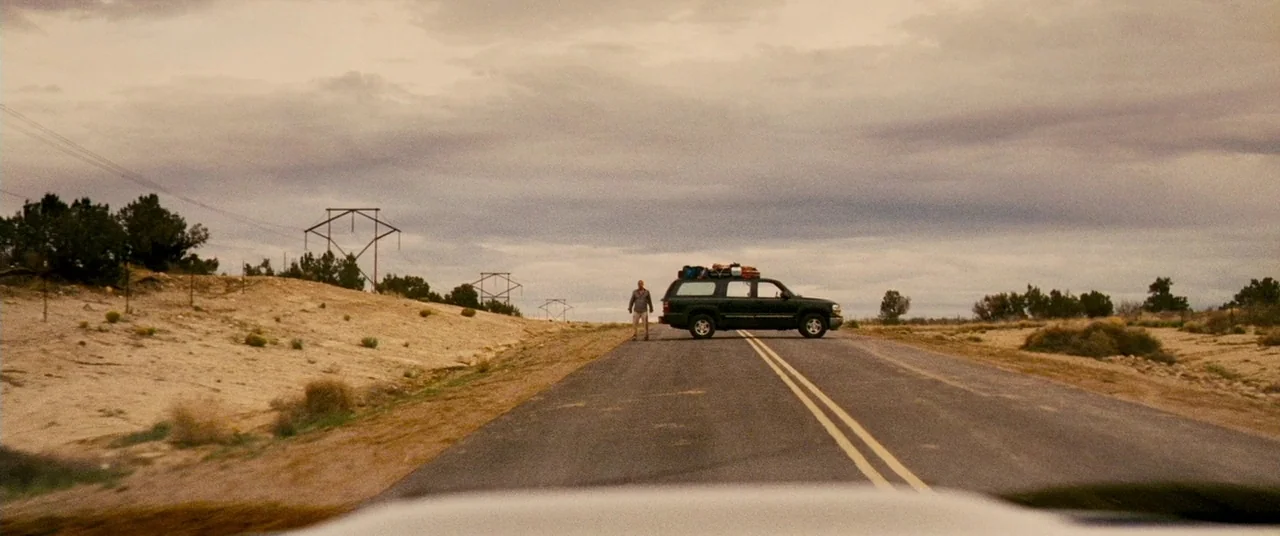 Carriers (2009) movie still: wide — A rural asphalt highway stretches through a flat, arid desert under a cloudy sky. An SUV with roof cargo is…