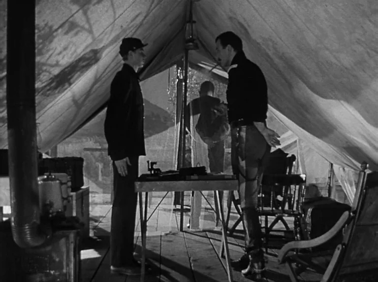 Rio Grande (1950) movie still: fullbody — A canvas military tent interior contains a wooden table and a metal stove. Two soldiers stand by the table…