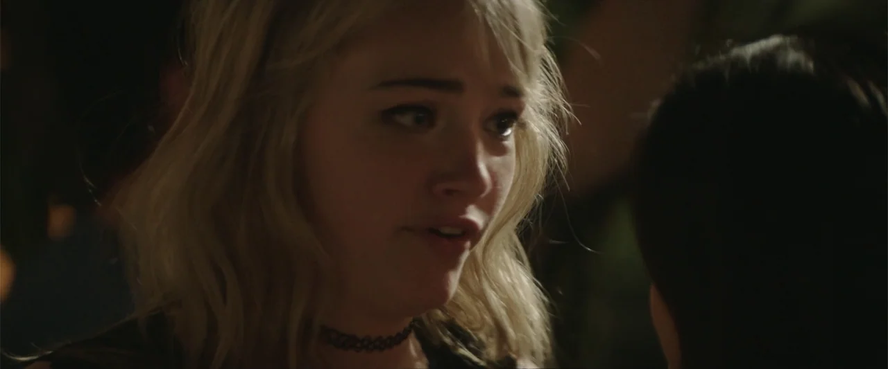 Truth or Dare (2018) movie still: closeup — A dimly lit indoor space with blurred background elements visible in the shadows. A young blonde woman…