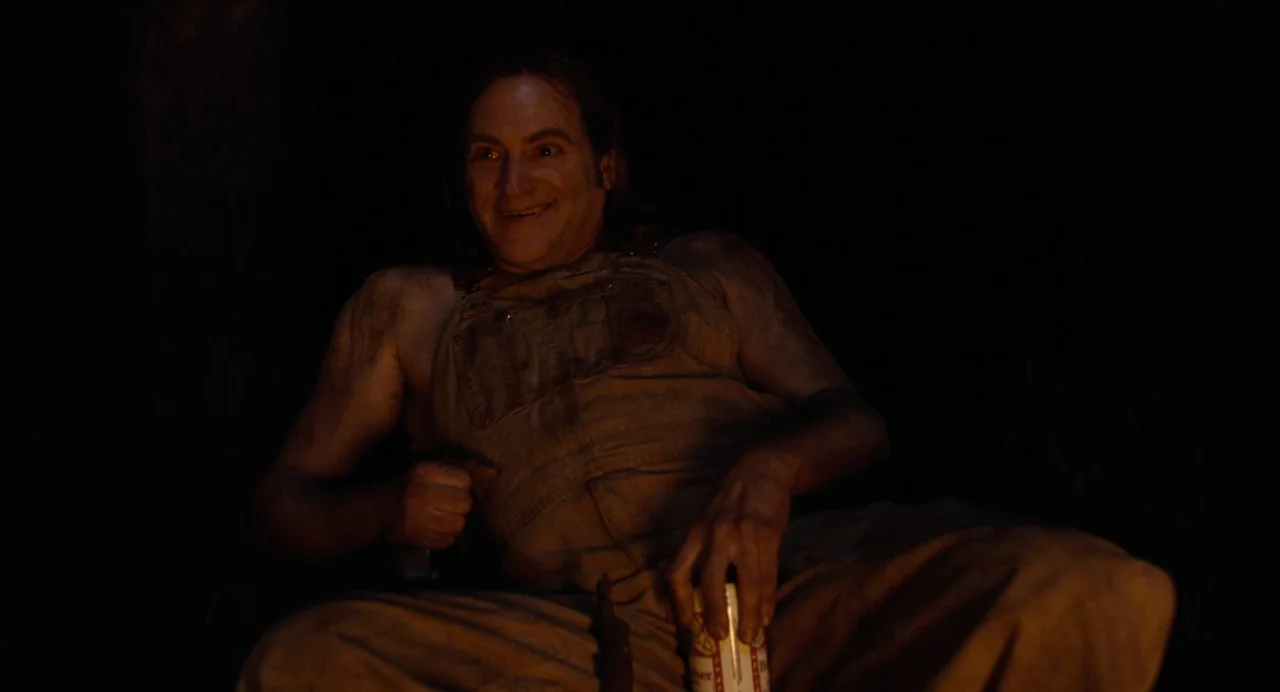 Bones and All (2022) movie still: medium — A man sits outdoors at night, illuminated by the low light of a fire. He is dressed in dirty work…