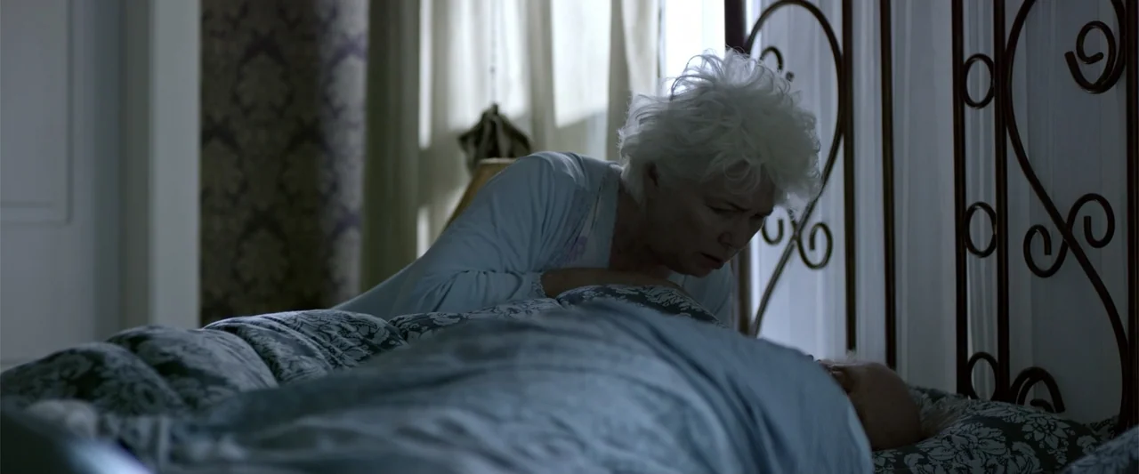 Dirty Little Secret (1998) movie still: medium — An interior bedroom features patterned wallpaper, sheer curtains, and a metal bed frame. An elderly…