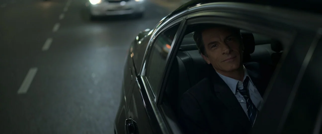 Doublespeak (2021) movie still: medium — A man sits in the back of a dark vehicle driving at night. He is turned to look directly at the viewer…