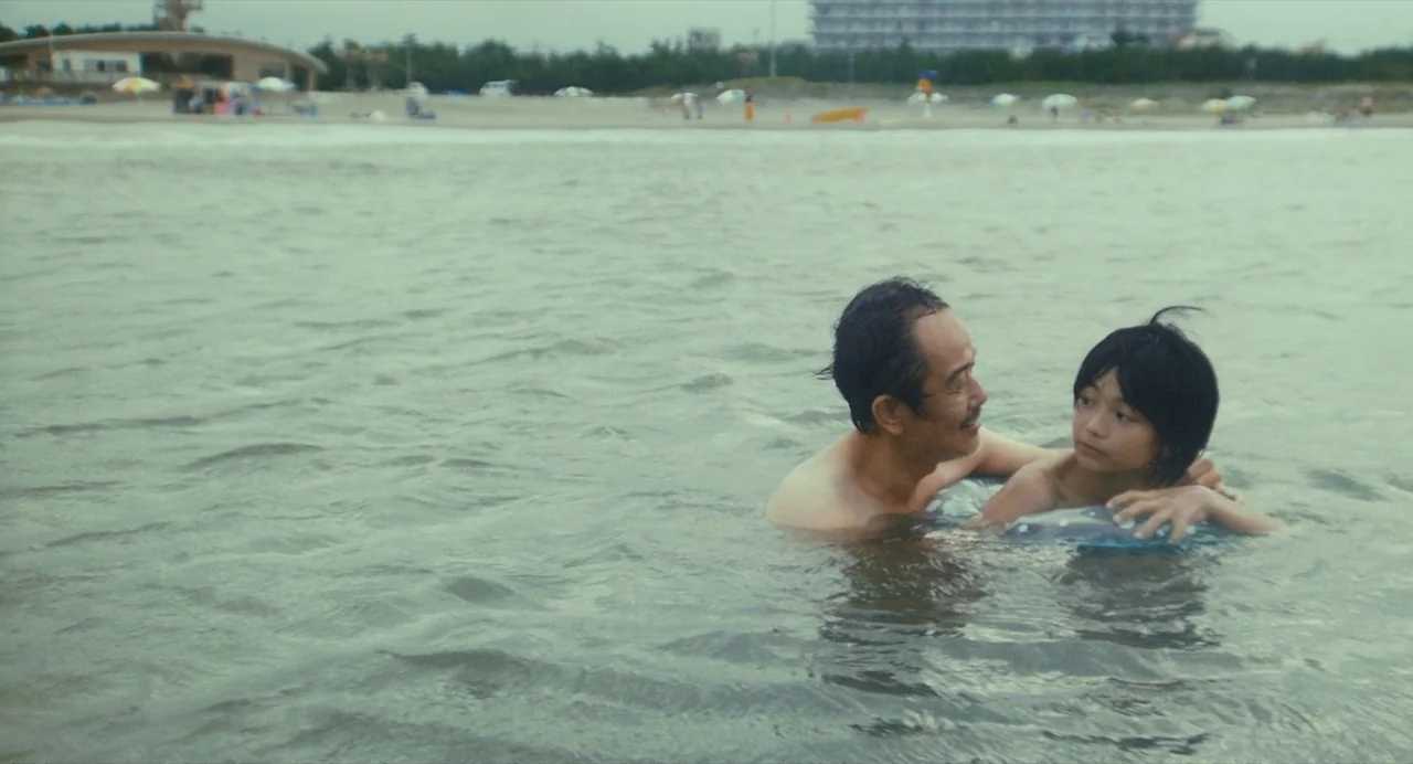 Shoplifters (2018) movie still: wide — A calm, grey ocean under an overcast sky with a distant sandy beach shoreline. An older man supports a young…
