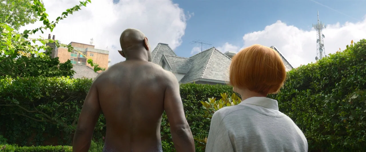 Three Thousand Years of Longing (2022) movie still: medium — A residential yard with dense green hedges and a view of a shingled roof. A muscular…