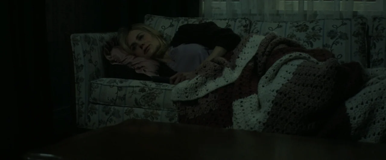 Holland (2025) movie still: medium — A dimly lit living room features a patterned fabric sofa with a crochet blanket. A woman in a dark sweater…