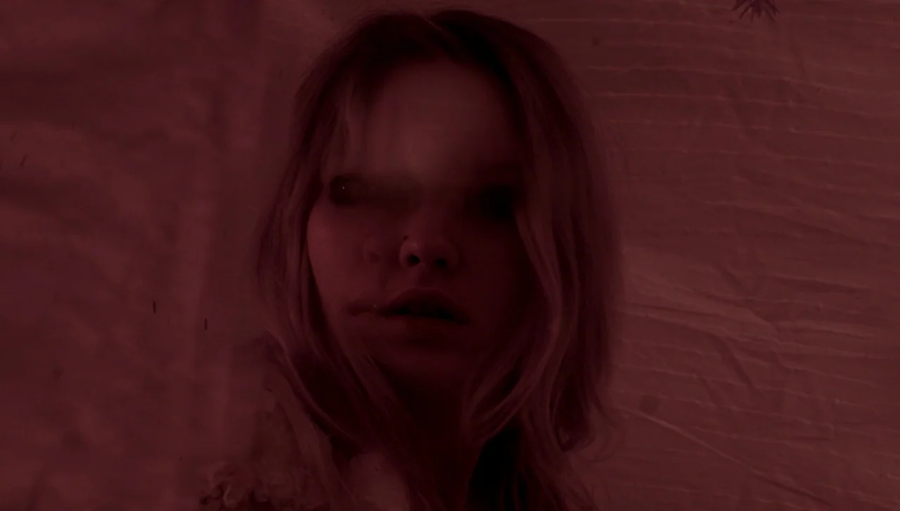 Hellions (2015) movie still: closeup — A person is captured in a dimly lit, monochromatic red environment with textured walls. The individual gazes…