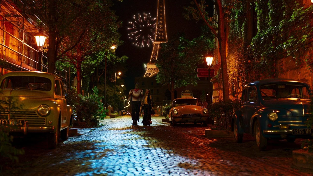The Munsters (2022) movie still: wide — A wet cobblestone street in Paris at night with two people walking toward camera. The Eiffel Tower is…