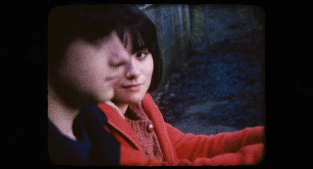 Submarine movie still: medium — An outdoor area at dusk with a dark fence in the background. Two young people sit closely together facing forward in…