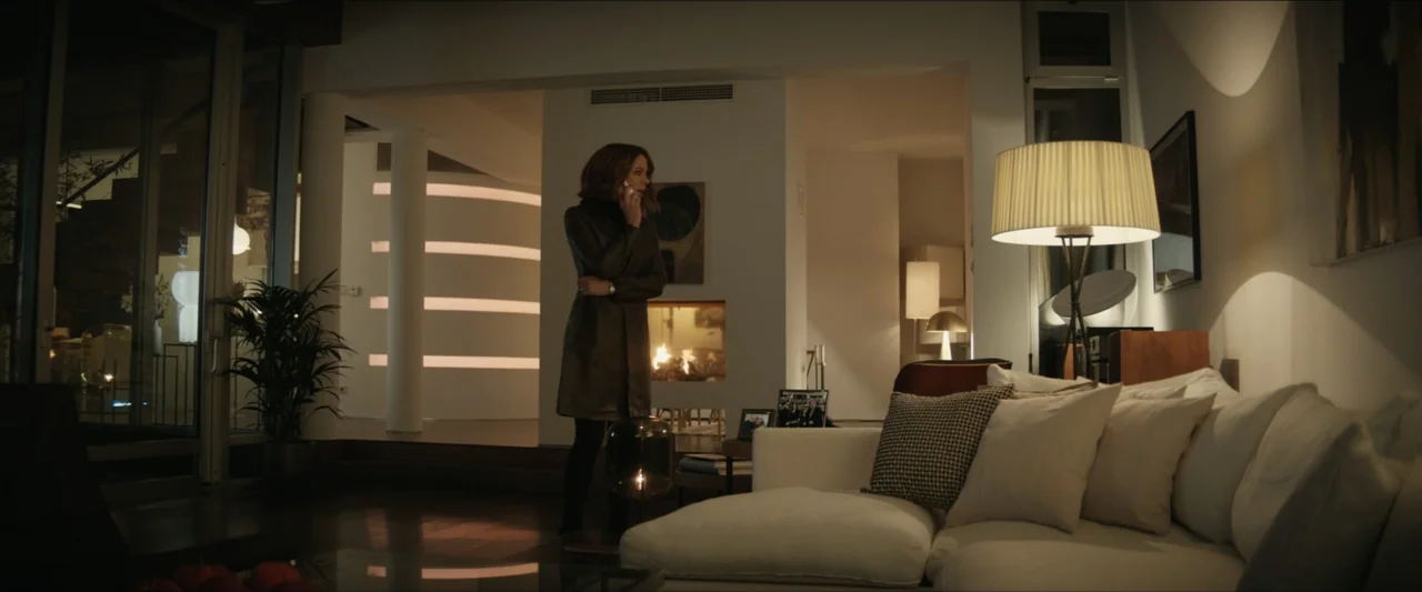 Canary Black (2024) movie still: fullbody — A spacious contemporary home interior features a white sofa, a fireplace, and ambient lighting. A woman…