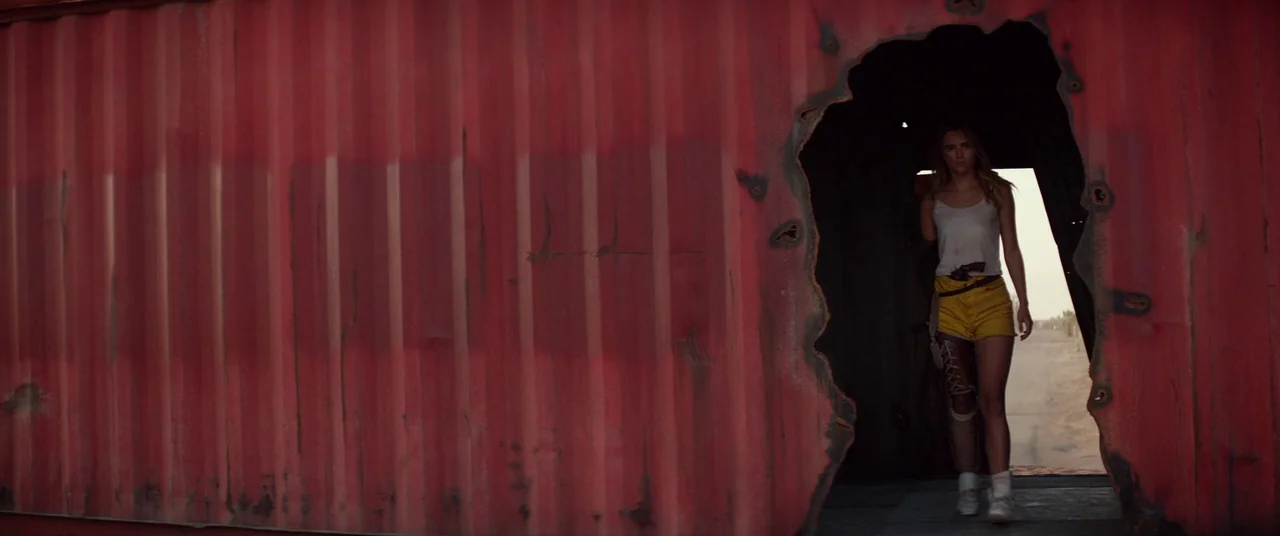 The Bad Batch (2016) movie still: wide — A rusted red metal shipping container features a large jagged hole cut out. A young woman walks through the…