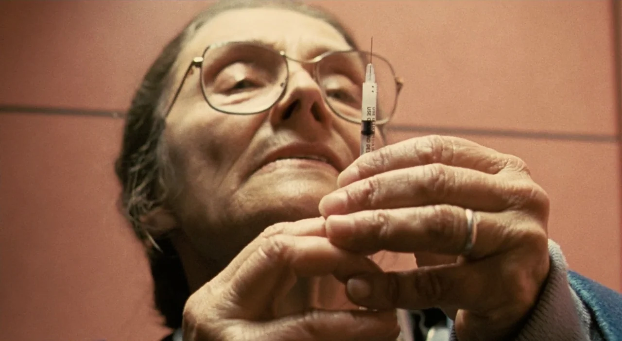 Patagonia (2010) movie still: closeup — An elderly woman stands before a flat, dull-colored wall in a clinical space. She holds a small medical…