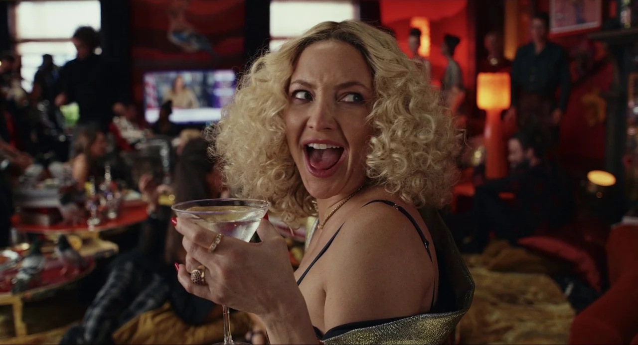 Glass Onion (2022) movie still: medium — A crowded social gathering space features red walls and dim, warm lighting accents. A blonde woman in a…