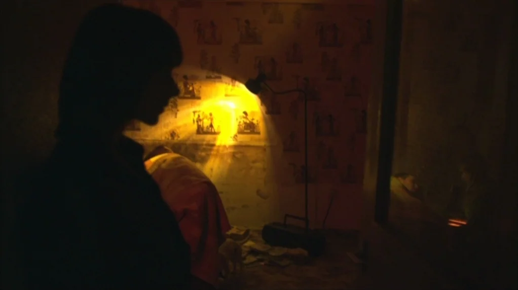 Red Road (2006) movie still: medium — A dark, dimly lit room features patterned wallpaper and a prominent yellow floor lamp. A person in shadow…
