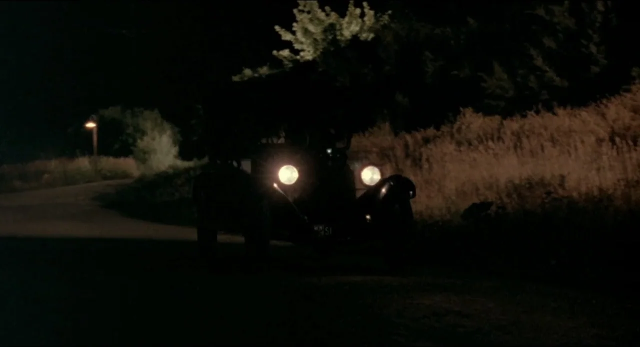 A River Runs Through It (1992) movie still: wide — A dark, unpaved country road is bordered by overgrown dry grass and trees. A vintage car with…
