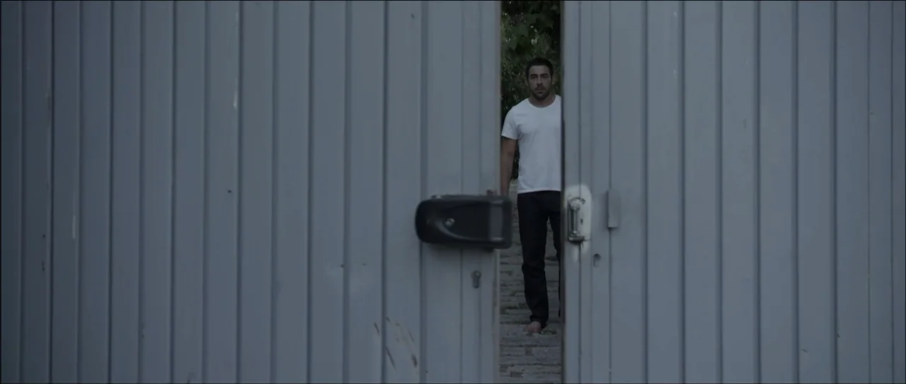 Otro verano (2013) movie still: medium — A man stands behind a half-open metal gate with vertical slats. He looks directly ahead through the narrow…