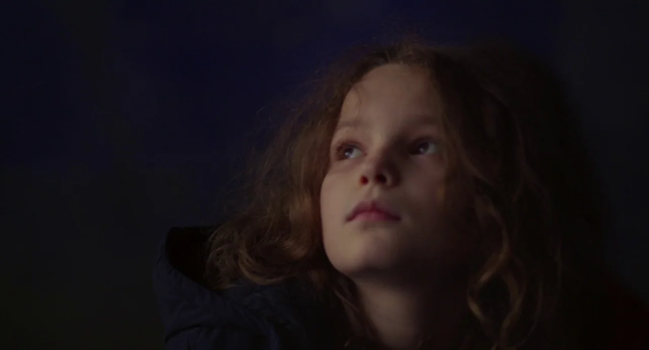 Petite Maman (2021) movie still: closeup — A dark, undifferentiated background frames a child in a dimly lit setting. The child tilts their head…