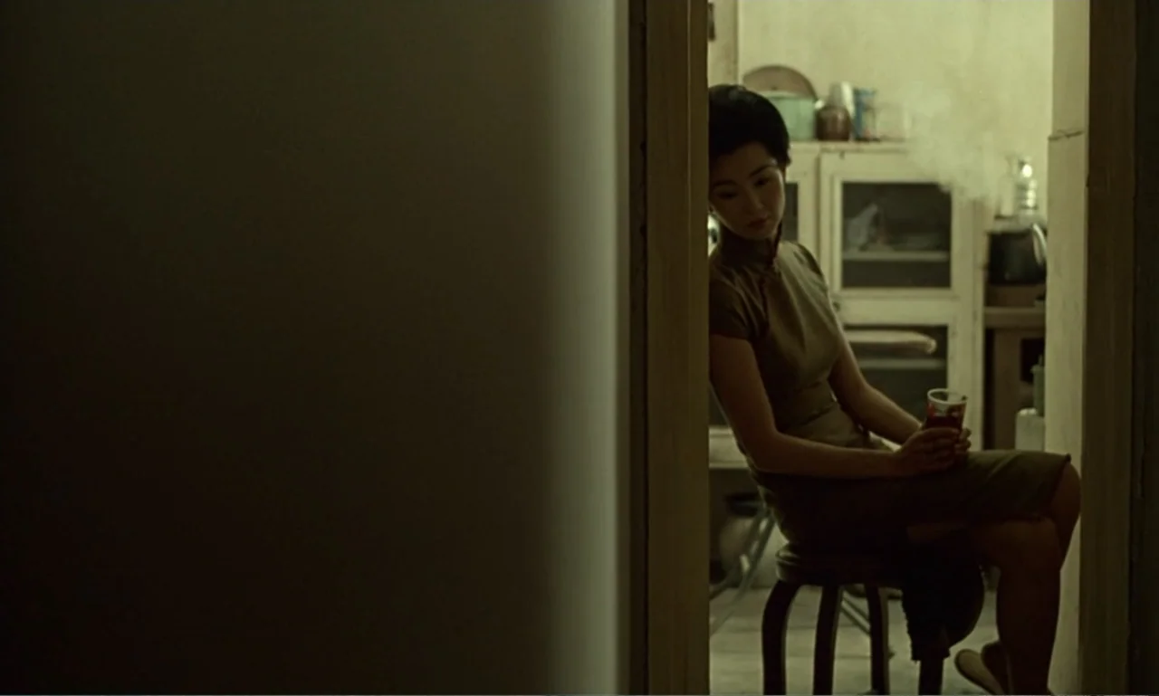 In the Mood for Love (2000) movie still: medium — A dimly lit room viewed through a narrow dark doorway. A woman in a traditional dress sits on a…