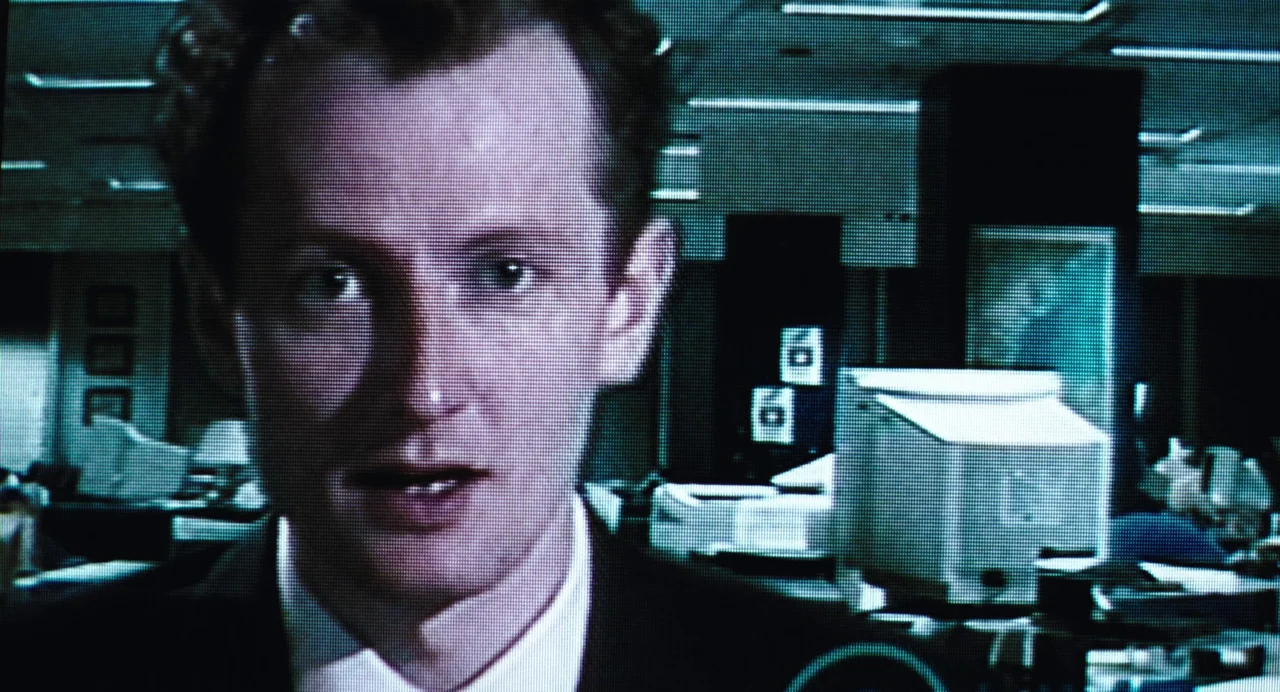 Last Night (1998) movie still: closeup — A dimly lit professional workspace contains cubicles, computer monitors, and ceiling-mounted fluorescent…