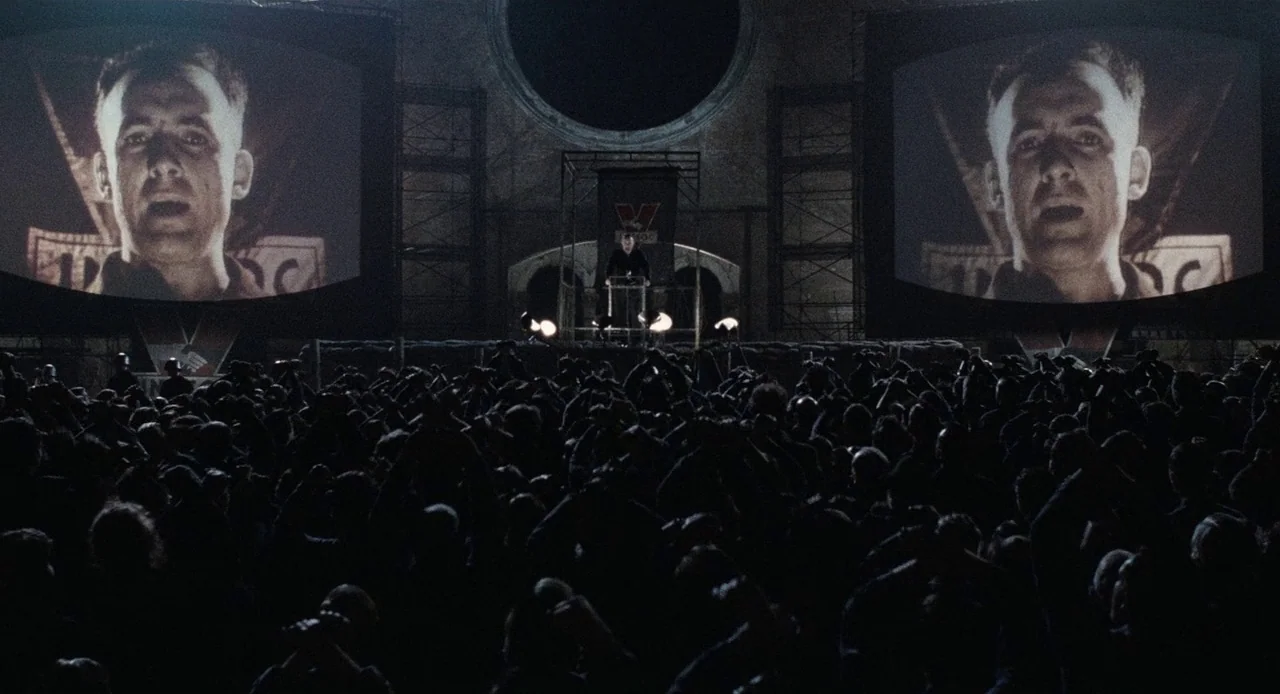 1984 (1984) movie still: wide — A massive crowd gathered in a dark, concrete-walled square looks at two large screens. On the screens, a man's face…