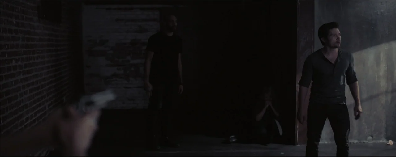 El Último Juego (2025) movie still: medium — A dimly lit industrial interior features a red brick wall and exposed concrete surfaces. Two men face…