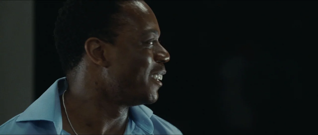Slackers (2002) movie still: closeup — A dark indoor setting with the subject positioned against a black backdrop. A man in a blue shirt is smiling…