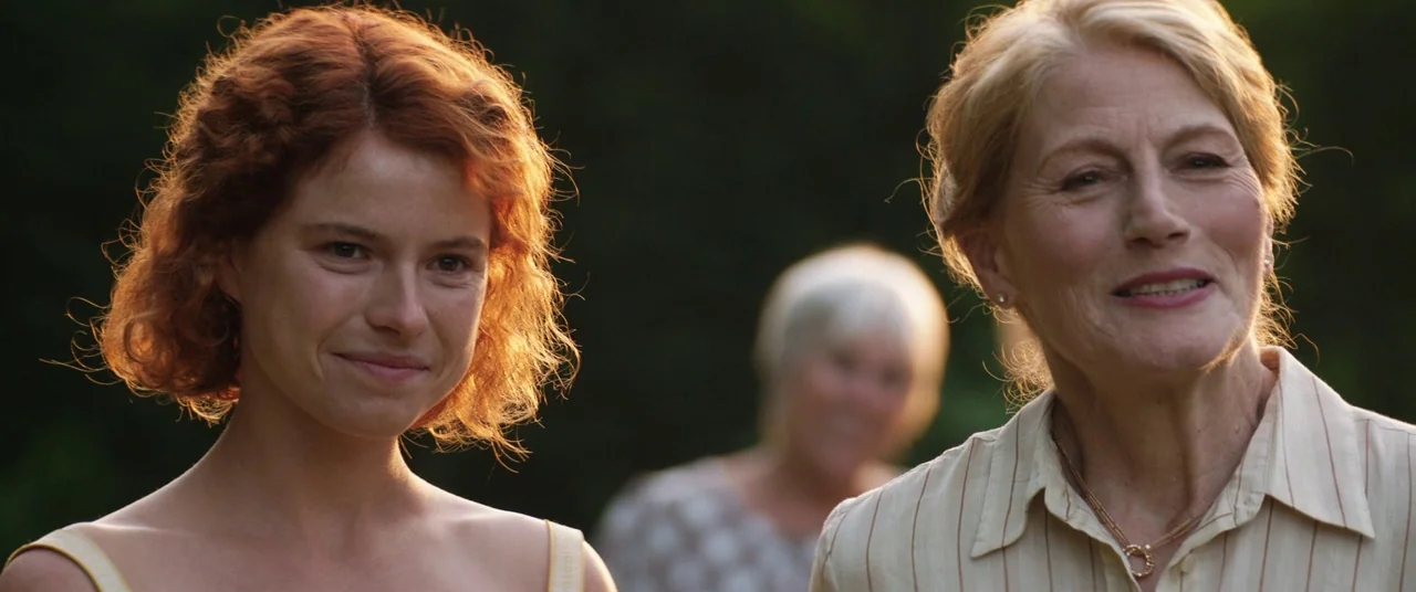Beast (2017) movie still: medium — Two women stand in an outdoor setting with blurred green foliage in the background. They are positioned…