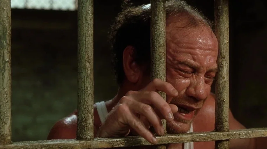 The Green Mile (1999) movie still: closeup — A dimly lit room features heavy vertical metal bars in the foreground. A middle-aged man with damp skin…