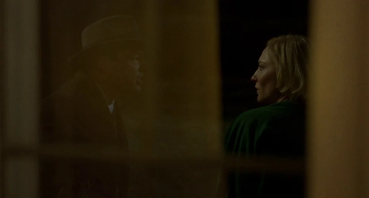 Carol (2015) movie still: medium — A dark, shadowed corridor with warm, low lighting reflecting off a glass partition. A man in a hat faces a woman…