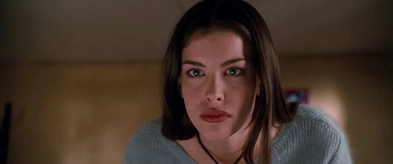Empire Records (1995) movie still: closeup — A dimly lit indoor space with a plain, beige wall background. A young woman looks directly toward the…