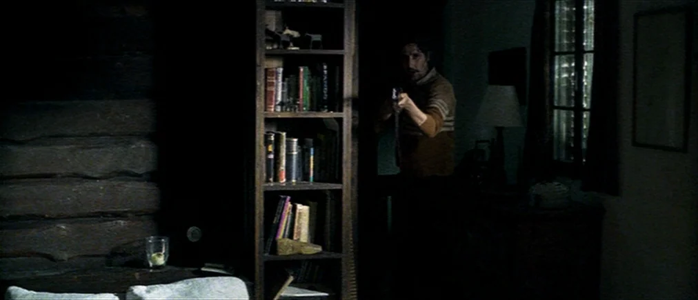 Time Of The Wolf (2003) movie still: medium — A dimly lit room with log walls features a tall bookshelf filled with various items. A man with a…