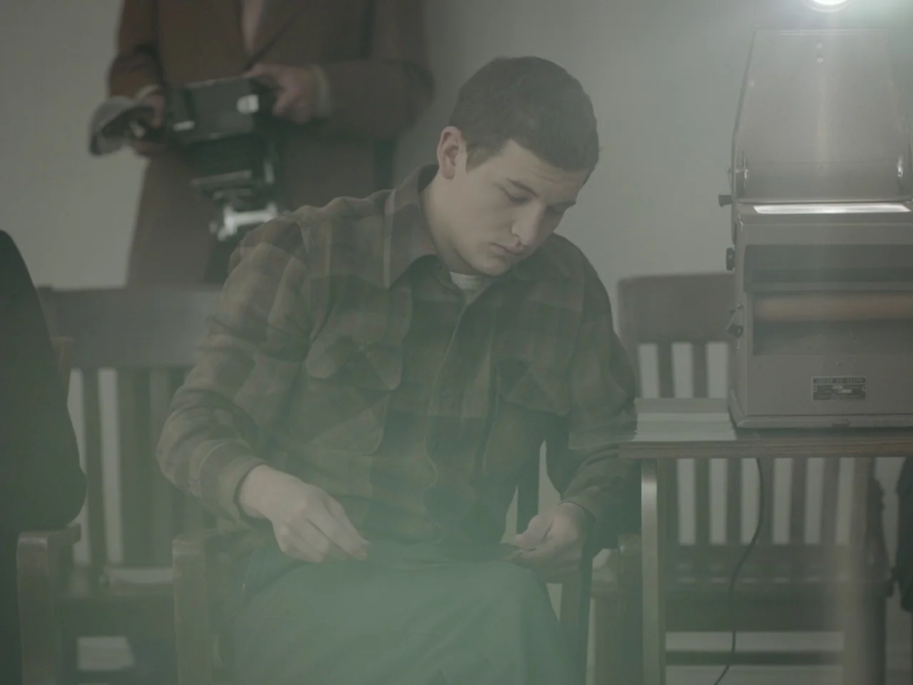 The Mountain (2018) movie still: medium — A muted indoor space features wooden chairs and a vintage overhead projector machine. A young man wearing…