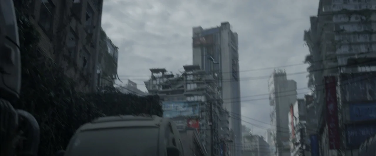 Godzilla (2014) movie still: establishing — A dense urban street shows damaged high-rise buildings under a heavy, overcast grey sky. A small…
