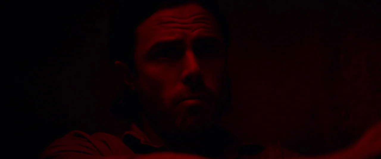 Triple 9 (2016) movie still: closeup — A dimly lit room is bathed in a saturated red light. A man stares forward from the shadows with a neutral…
