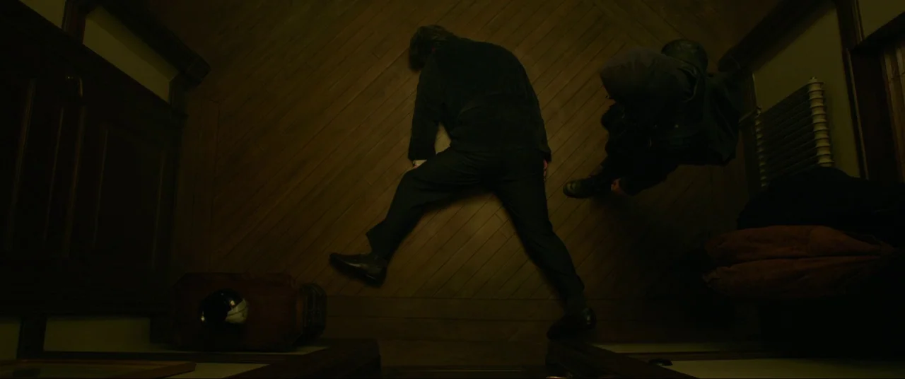 The Girl With The Dragon Tattoo (2011) movie still: wide — A dimly lit narrow hallway has hardwood flooring and dark wood trim details. A man lies…