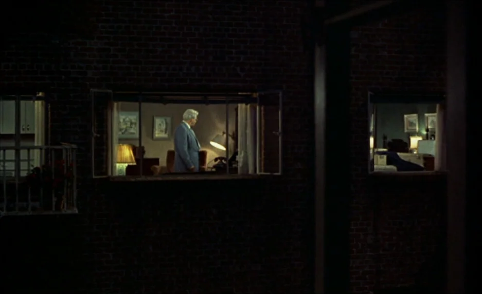 Rear Window (1954) movie still: wide — A dark brick apartment building at night reveals illuminated interiors through two framed windows. An older…