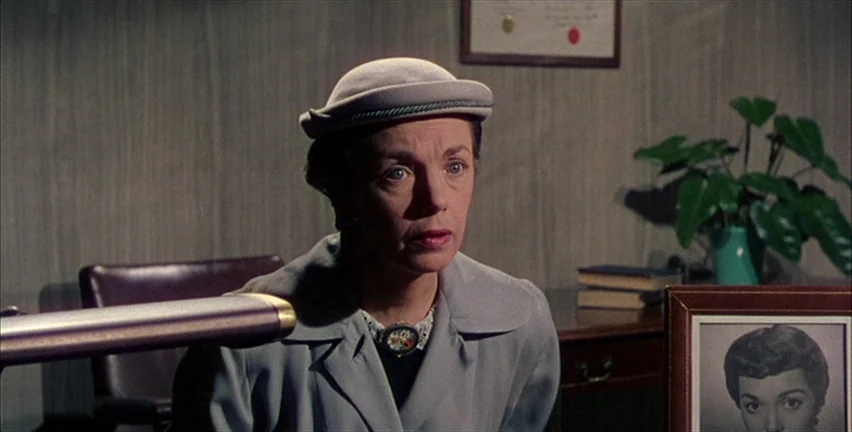 Magnificent Obsession (1954) movie still: medium — A quiet room features wood-paneled walls and a desk with a plant. A woman in a pillbox hat sits…
