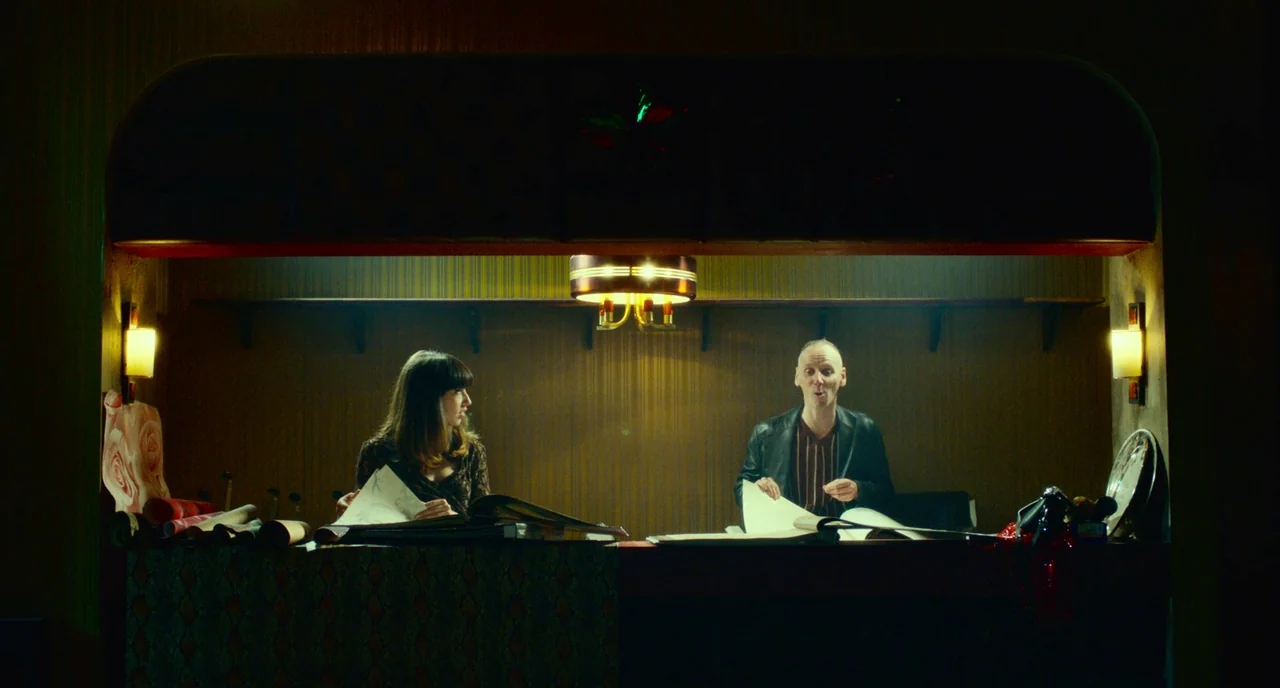 T2 Trainspotting (2017) movie still: medium — A dimly lit room features a service counter with vertical wood-paneled walls and wallpaper. A woman…